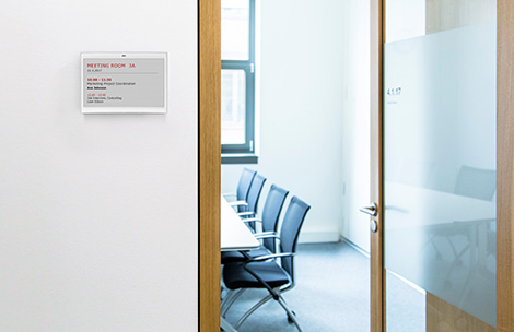 Empty meeting room with open door. On the wall on the left hand side, there has been mounted a LANCOM Wireless ePaper, displaying the next meeting.