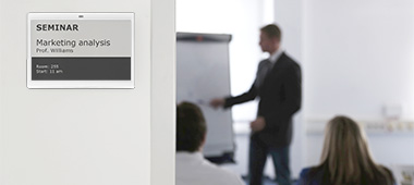 Signage of a seminar room by means of a LANCOM Wireless ePaper Display. A young man wearing a suit is giving a presentation.
